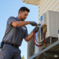 HVAC technician installing an outdoor unit, carefully connecting refrigerant lines with tools. The professional is wearing a grey uniform shirt and work gloves, demonstrating precision and expertise on a sunny day.